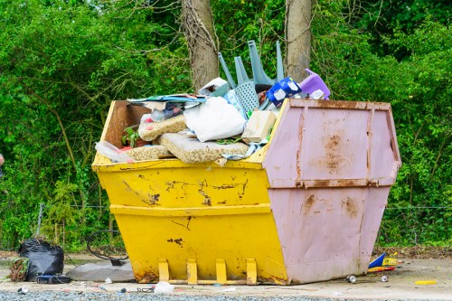 A large open-top skip bin positioned on a gravel surface outdoors, filled with a variety of domestic rubbish including plastic storage containers, foam packaging, cardboard boxes, and used cleaning supplies. The bin has a yellow base with a pinkish-burple upper section, both showing signs of dirt and rust. Behind the skip, there are tall trees and green foliage, indicating a natural outdoor setting, possibly near a residential area or garden. To the left of the bin, a black rubbish bag rests on the gravel, and the surrounding environment appears to be a collection point for waste disposal, consistent with professional rubbish removal services such as those offered by Garden Clearance Walthamstow in the London area.