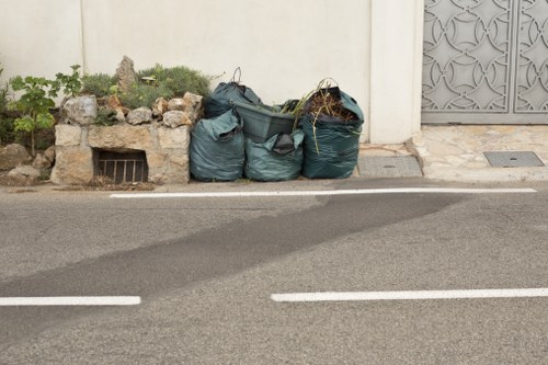 The image shows three large green construction rubbish bags filled with waste, placed on the edge of a paved street curb. The bags appear to contain mixed materials, including some visible branches or plant waste, with the bags leaning slightly against a low stone wall that has some greenery and small plants growing near it. Behind the bags is a plain, off-white wall, and to the right, part of an ornate metal gate is visible, suggesting a residential property. The street surface is dark grey asphalt with white road markings, and the scene is illuminated by natural daylight, indicating a clear or partly cloudy day. This setting reflects a typical rubbish collection or clearance scene, in a neighbourhood where Garden Clearance Walthamstow operates, subtly connecting the visual to local waste removal services for garden and general rubbish disposal in Walthamstow or the surrounding postcode area.