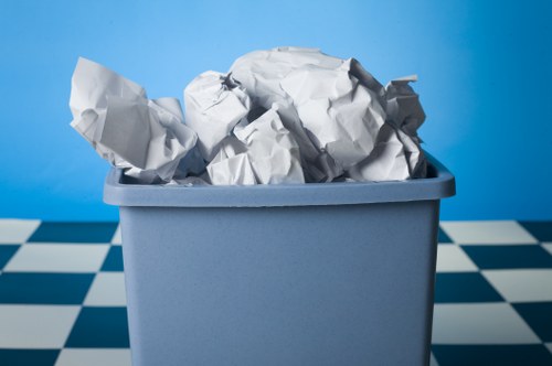 A large, light grey plastic waste bin filled with crumpled white paper and tissue paper, placed on a checkered black and white tiled floor. The background features a plain blue wall, suggesting an indoor or sheltered environment. The crumpled paper appears to be ordinary household waste, with the papers varying in size and shape, some with visible creases and folds. The bin is positioned centrally in the image, with the top edge of the container visible and the waste overflowing slightly. The scene captured by Garden Clearance Walthamstow illustrates typical rubbish disposal, relevant to rubbish removal services in Walthamstow involved in collecting and recycling household waste such as paper and tissue waste materials.